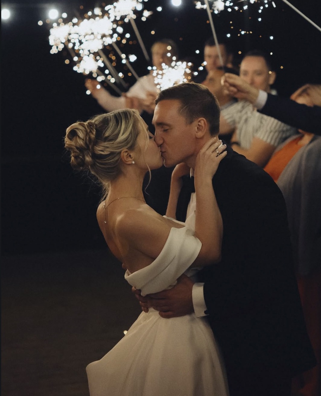 A kiss under sparklers at night: the bride in a white slip dress, the groom in black, guests holding lit sparklers around them