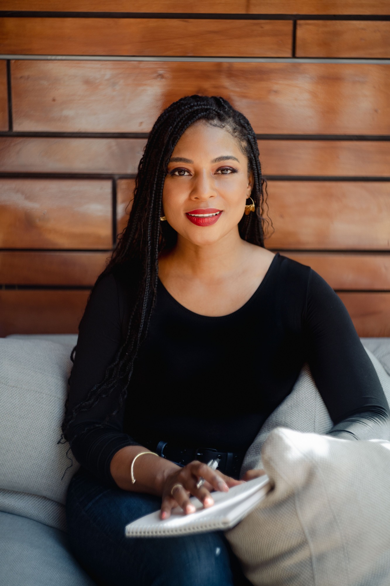 Personal-branding portrait: a woman with long braids and gold hoop earrings sits on a grey sofa with an open notebook, wood-panelled wall behind her