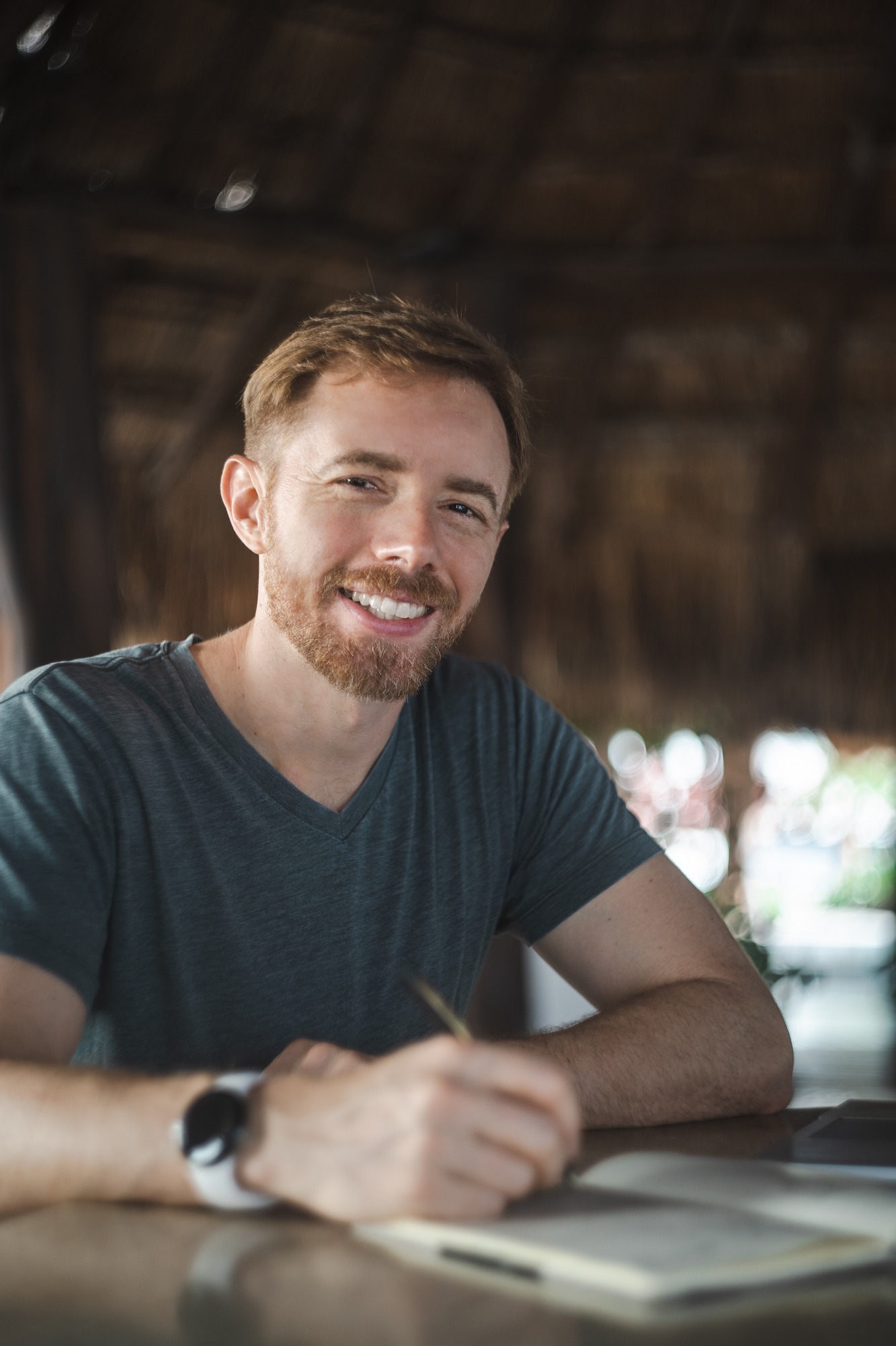 Portrait of a man with an auburn beard writing in a notebook at a table under a thatched palapa roof
