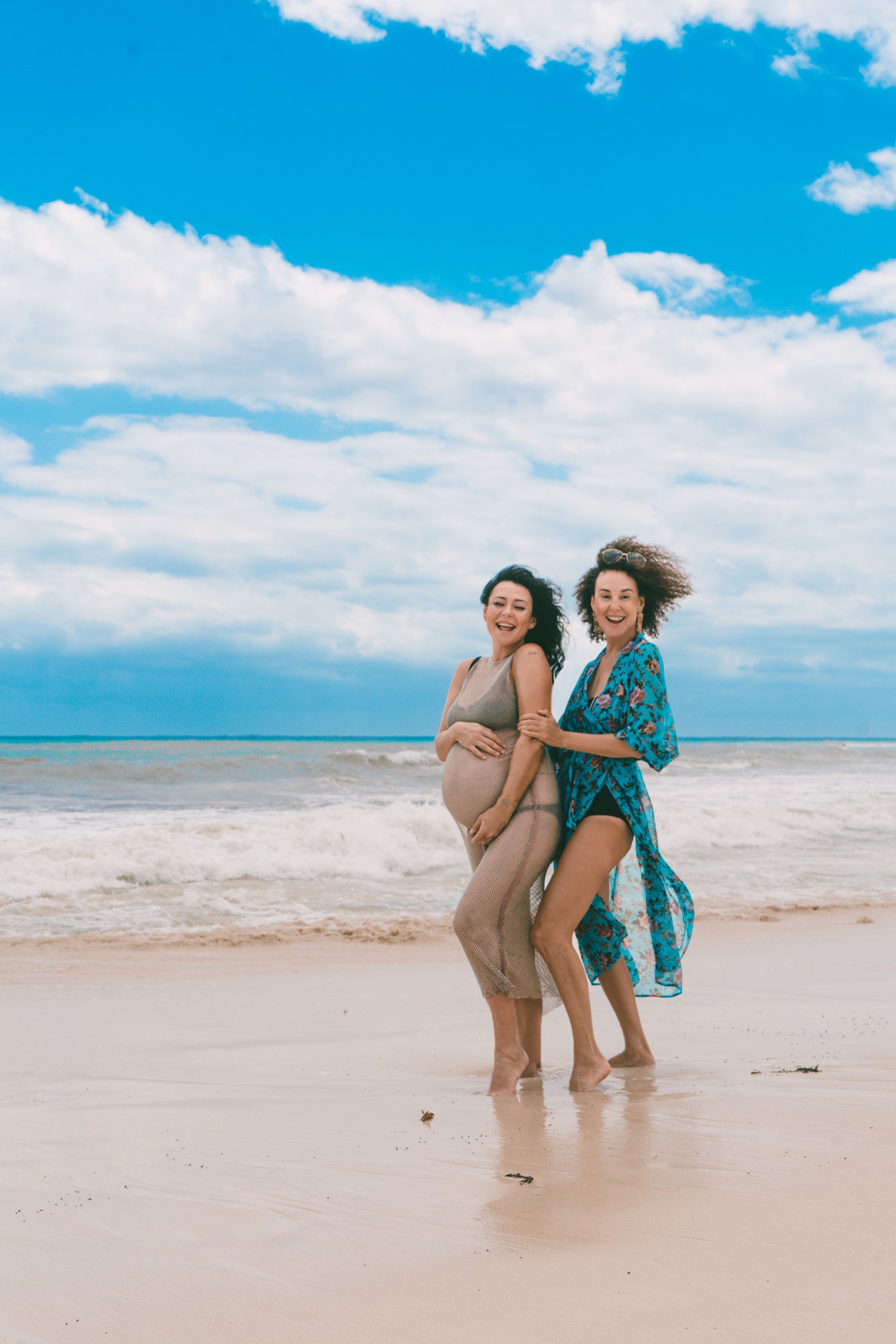 Maternity session on the beach: an expectant mother in a sheer beige dress laughs with a friend in a turquoise floral robe, surf and clouds behind them