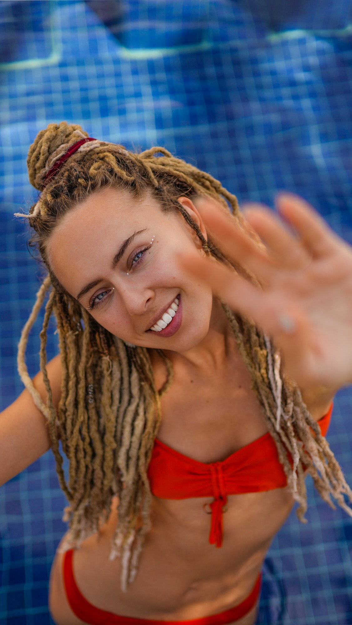 Young woman with blonde dreadlocks in a red bikini laughing in a blue-tiled pool, one hand reaching towards the camera